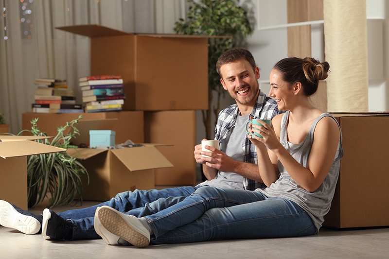 Happy young couple sitting on the floor, drinking coffee and smiling, with moving boxes around after a move.