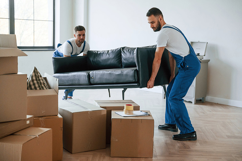 " Two professional movers moving a sofa with boxes and packing materials on the floor."